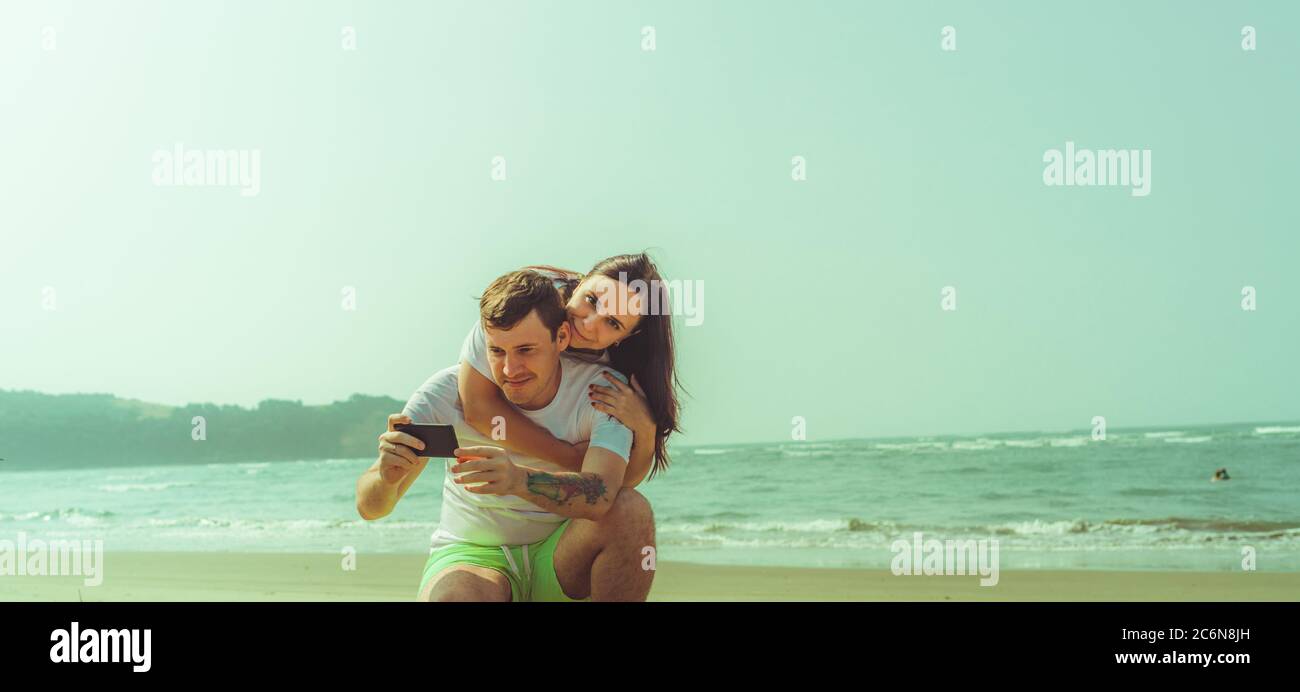 Happy couple taking selfies near sea. Loving couple embracing during date on beach against waving sea and cloudless sky. Stock Photo