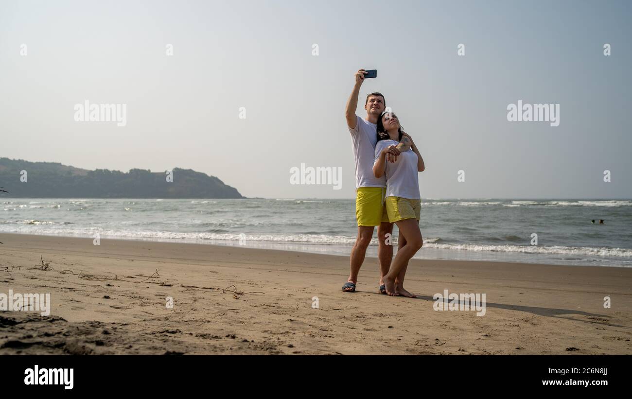 Happy couple taking selfies near sea. Loving couple embracing during date on beach against waving sea and cloudless sky. Stock Photo