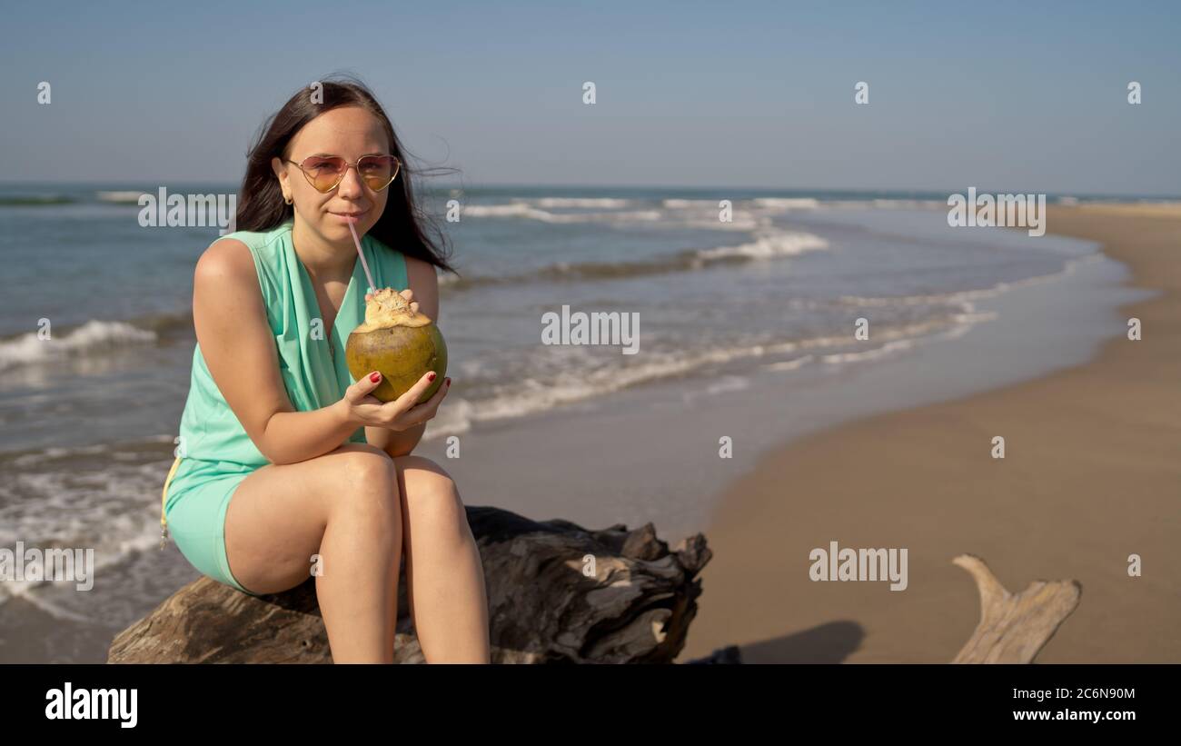 A beautiful young woman is sitting on a log and drinking the coconut by the sea or ocean in a bright sunny day. Stock Photo