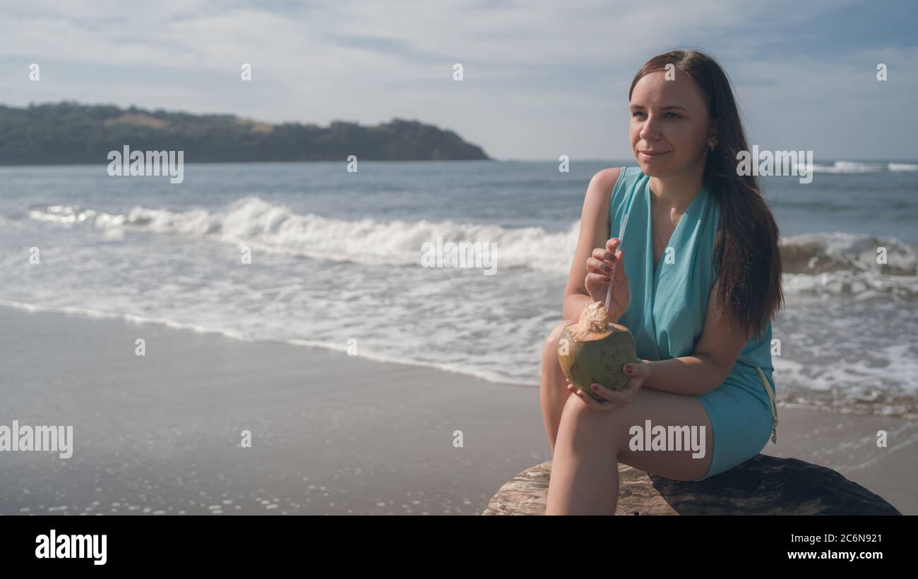 A beautiful young woman is sitting on a log and drinking the coconut by the sea or ocean in a bright sunny day. Stock Photo