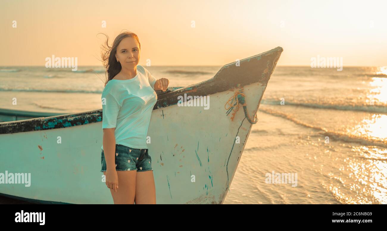 A young woman stands near the boat by the sea against the sunset. Stock Photo