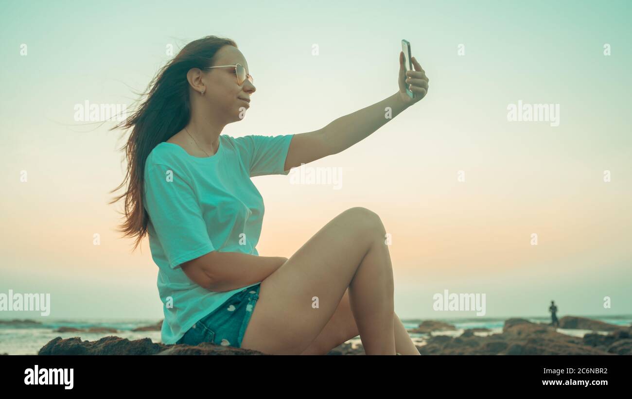 A beautiful young woman is taking selfie on a mobile phone, sitting on the rocks by the sea against the sunset. Stock Photo
