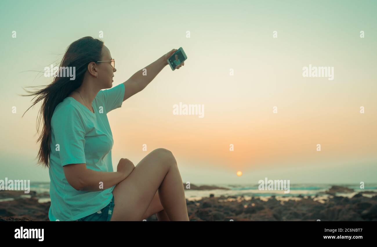 A beautiful young woman is taking selfie on a mobile phone, sitting on the rocks by the sea against the sunset. Stock Photo