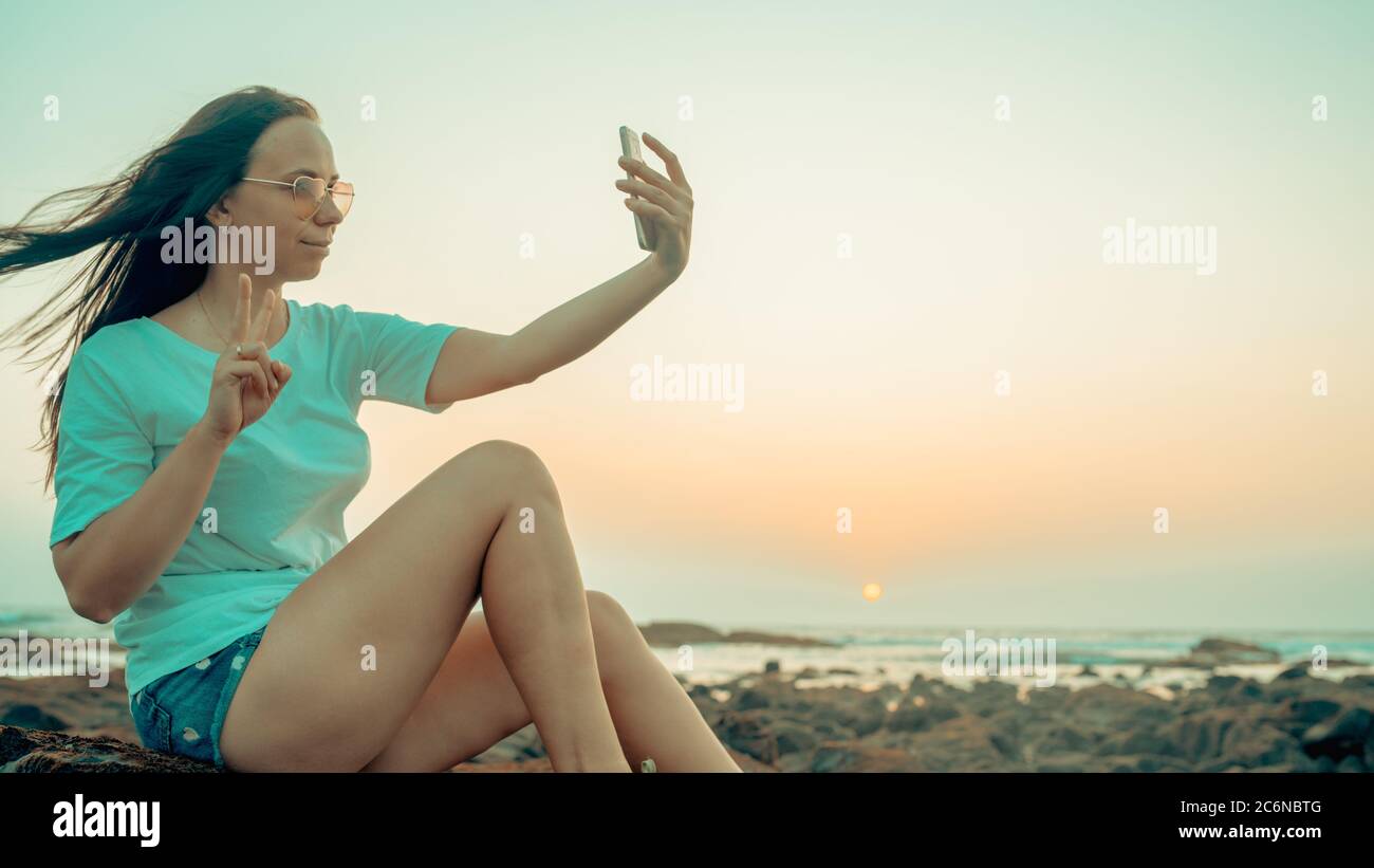 A beautiful young woman is taking selfie on a mobile phone, sitting on the rocks by the sea against the sunset. Stock Photo