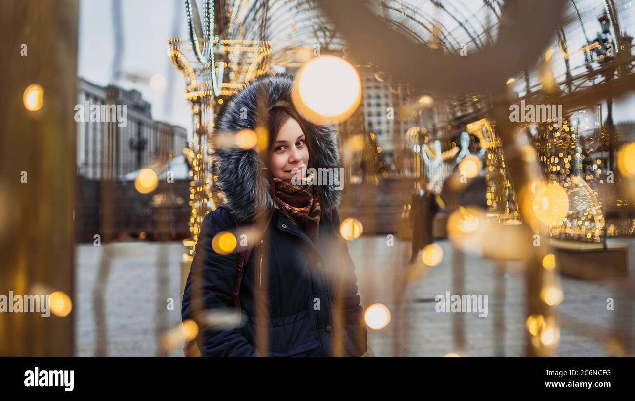 Portrait of a beautiful young woman in a jacket and hood through a garland on the street. Stock Photo