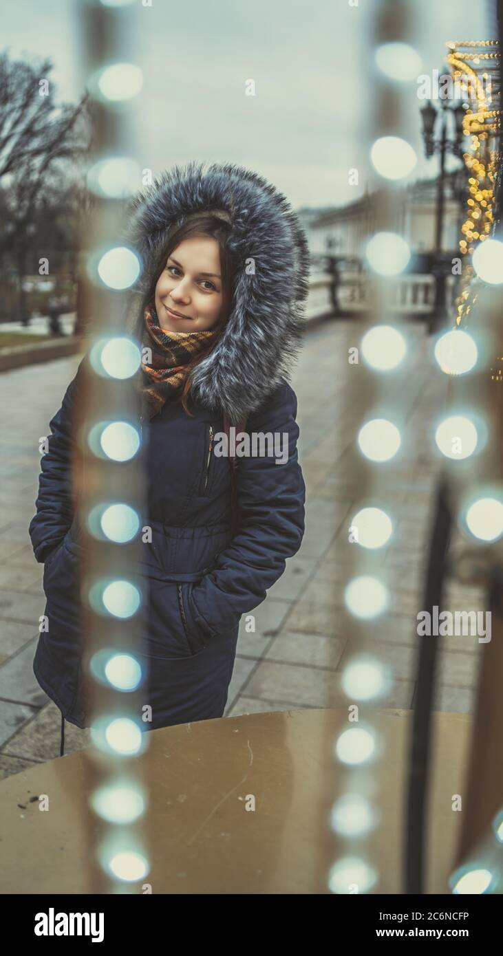 Portrait of a beautiful young woman in a jacket and hood through a garland on the street. Stock Photo