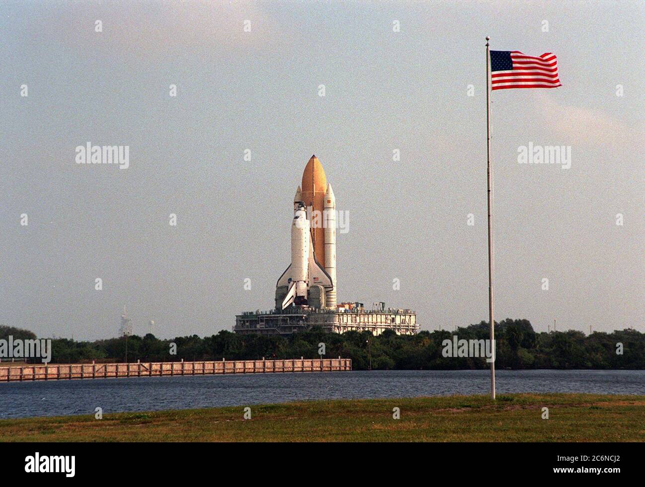 With the American flag flapping in the morning breeze, Space Shuttle Discovery, across the turn basin, makes its crawl to Launch Pad 39B (background, left) atop the mobile launcher platform and crawler transporter. Once at the pad, the orbiter, external tank and solid rocket boosters will undergo final preparations for the STS-103 launch targeted for Dec. 6, 1999, at 2:37 a.m. EST. Stock Photo