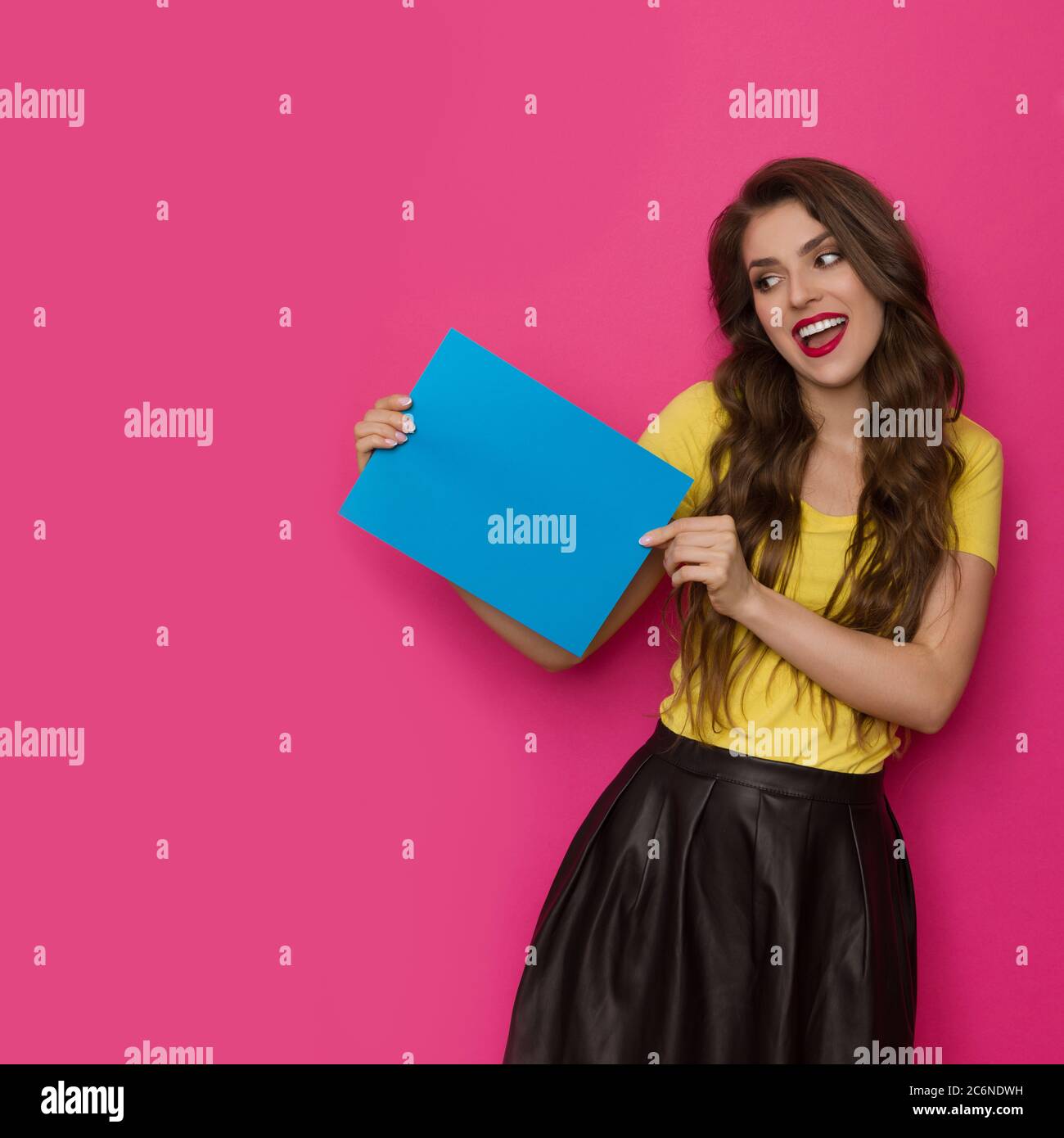 Happy beautiful young woman is holding empty blue sheet of paper and reading. Front view. Three quarter length studio shot on pink background. Stock Photo