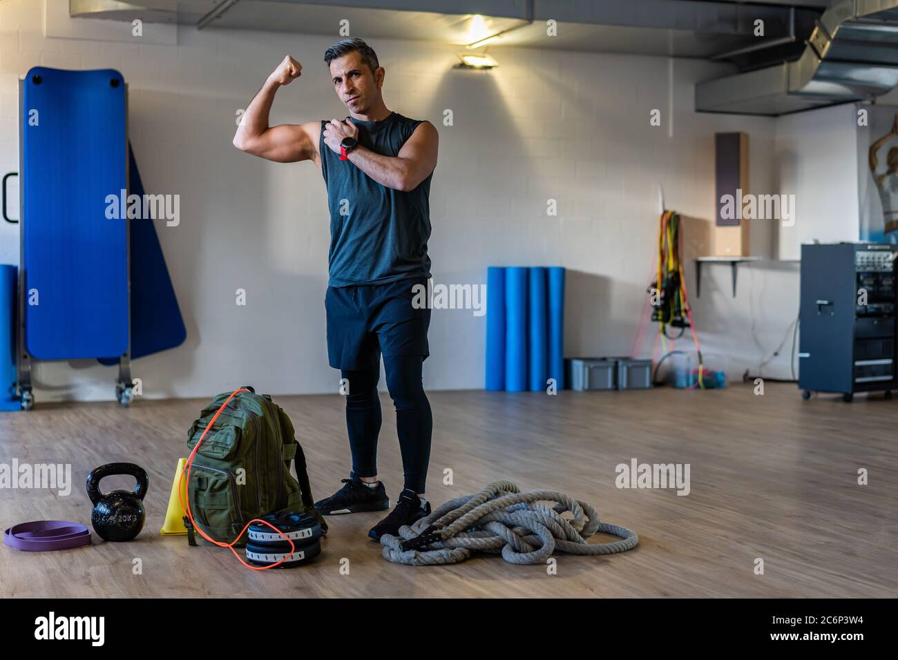 Motivated boot camp instructor stands with gym equipment in gym hall. Dumbbells, rope, sandbag on wooden floor. Portrait of instructor showing muscles Stock Photo