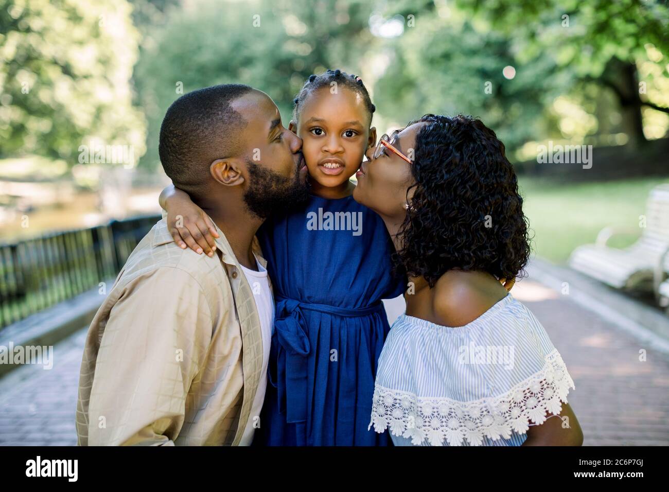 Adorable summer portrait of young african family of three posing together outdoors in beautiful park. Lovely parents are kissing their little daughter Stock Photo