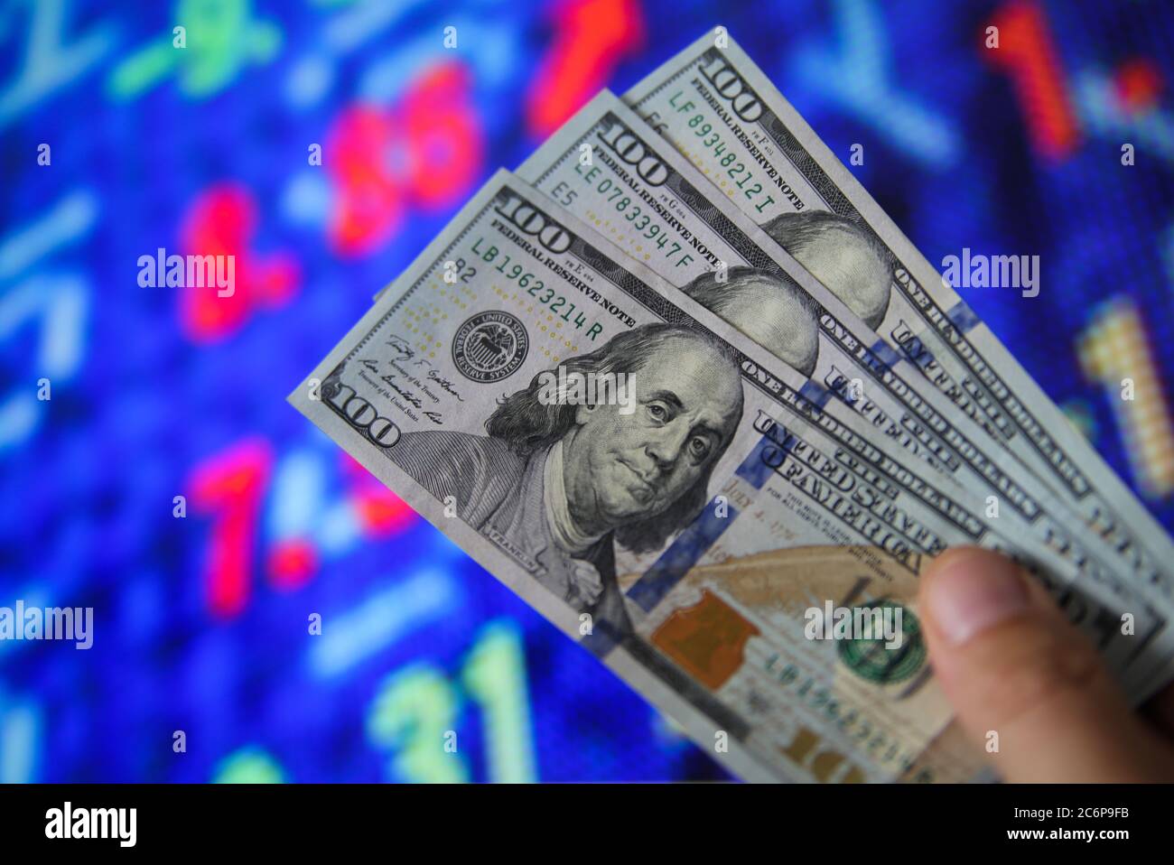 Investment withdrawal concept: View on hand holding three 100 dollar notes, blurred stock market exchange board background. (Focus on face) Stock Photo