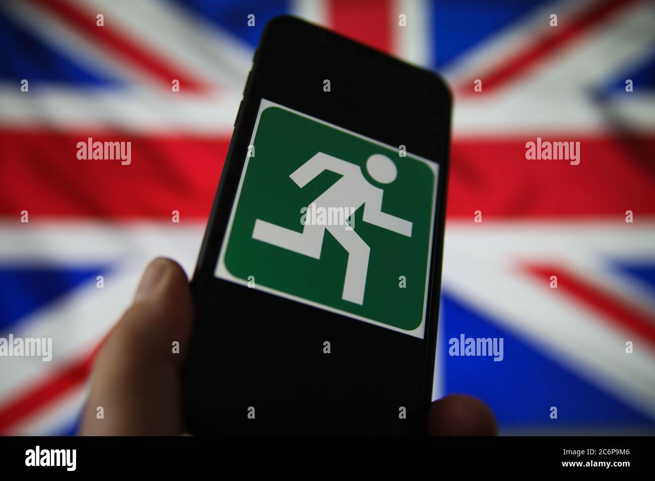 Viersen, Germany - July 9. 2020: View on isolated mobile phone screen with green emergency evacuation exit sign. Blurred union jack background. Stock Photo