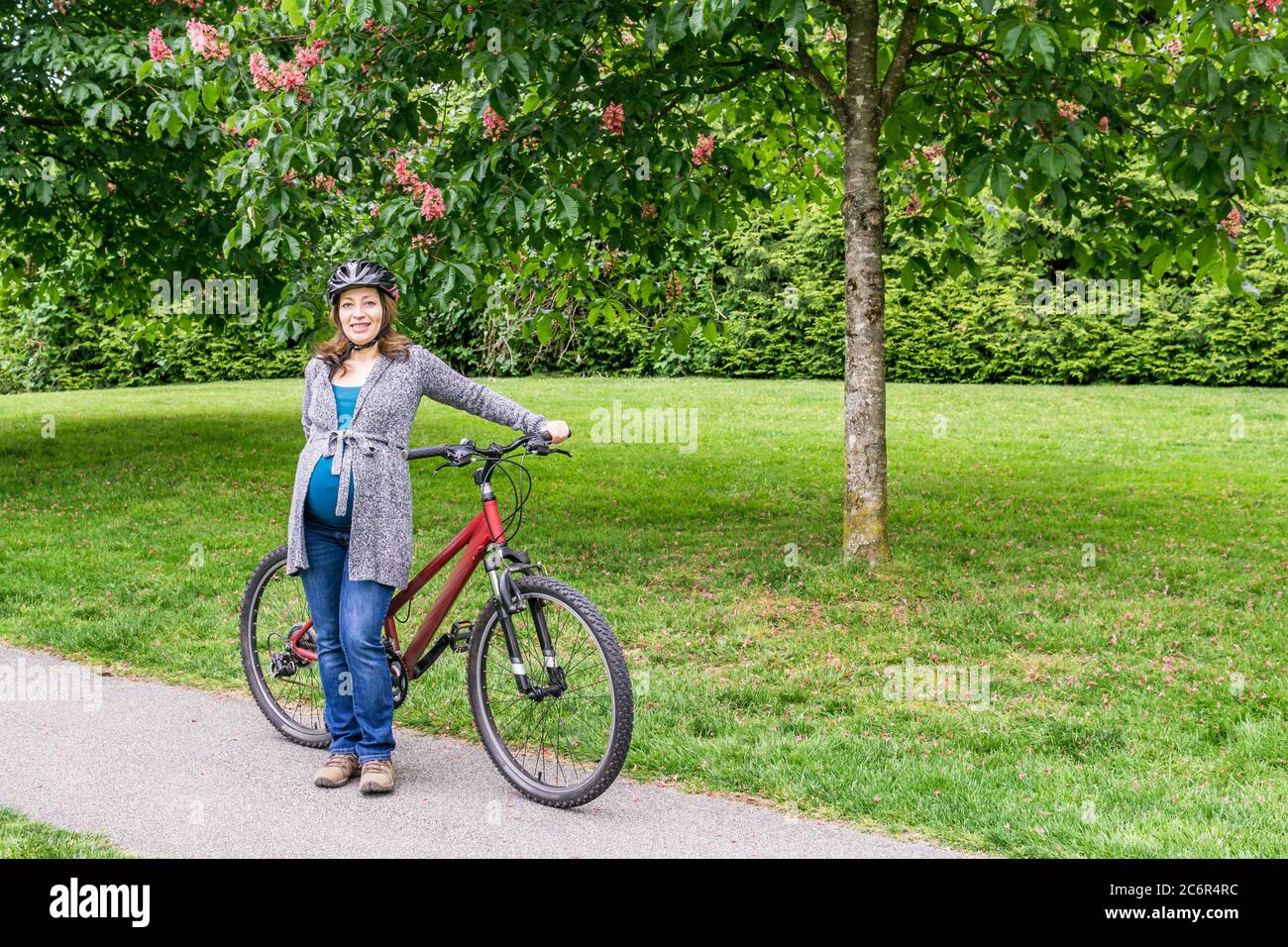 pregnant woman on the green lawn in a park holding bicycle ready to ride. Stock Photo