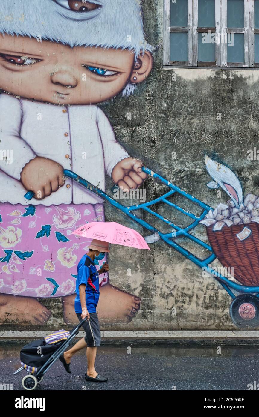 In the market in Phuket Town, Thailand, a man with umbrella passes a mural with three-eyed child character named Mardi by Thai street artist Alex Face Stock Photo