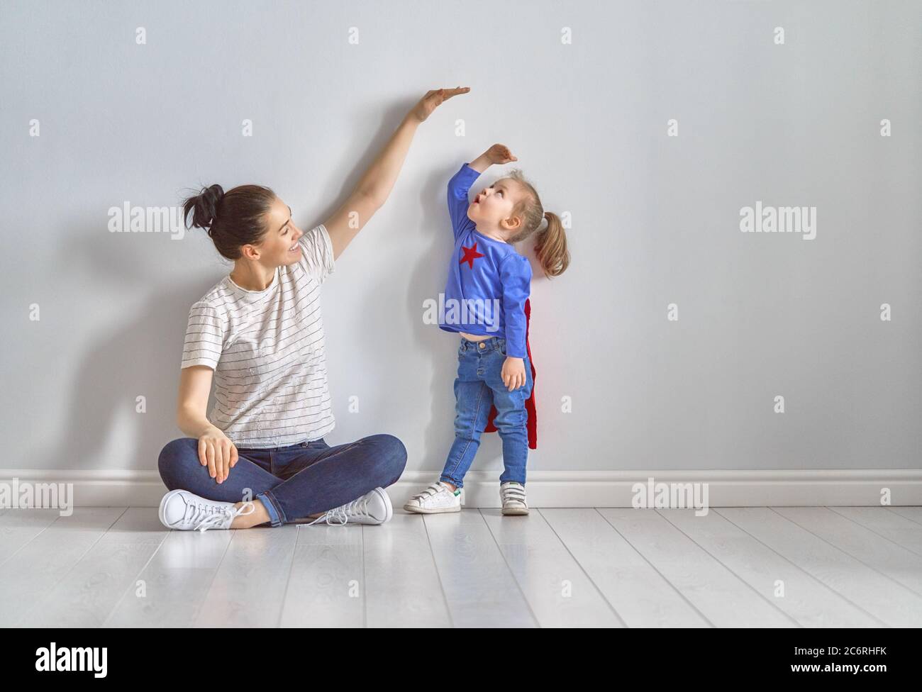 Mother is measuring growth of child daughter near empty wall. Girl in superhero costume. Stock Photo