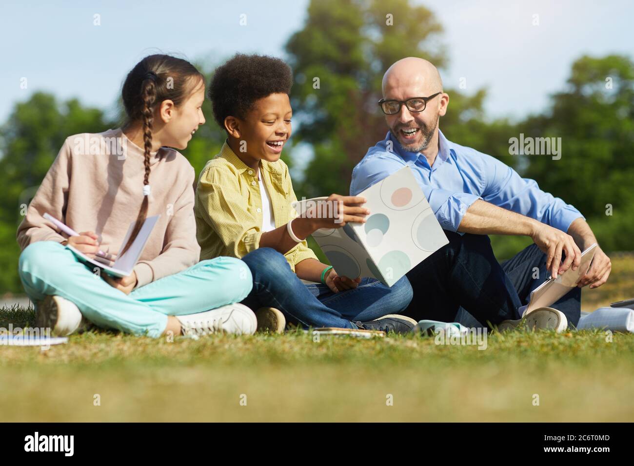 Full length portrait of smiling male teacher talking to children while sitting on green grass and enjoying outdoor class in sunlight Stock Photo