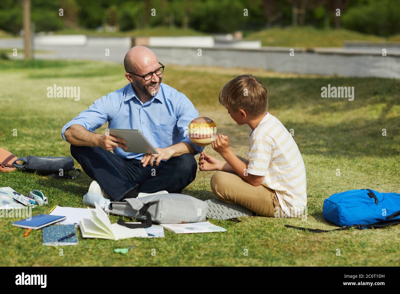 Full length portrait of smiling bald teacher talking to teenage boy holding model planet while enjoying outdoor astronomy lesson in sunlight, copy spa Stock Photo