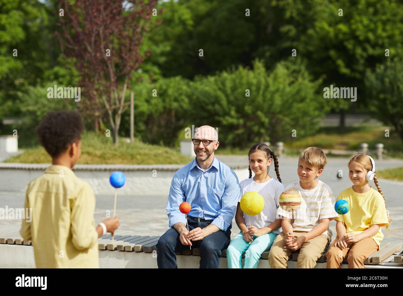 Portrait of smiling male teacher listening to boy giving presentation on astronomy while sitting with group of children and enjoying outdoor lesson in Stock Photo