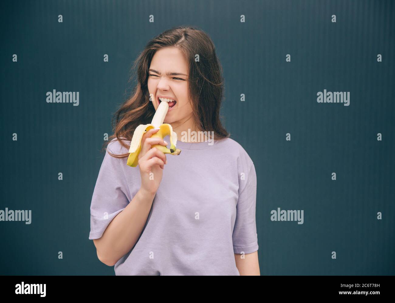 Young stylish trendy woman isolated over grey blue background. Girl biting piece of yellow ripe banana and doing weird facial expression. Stand alone Stock Photo