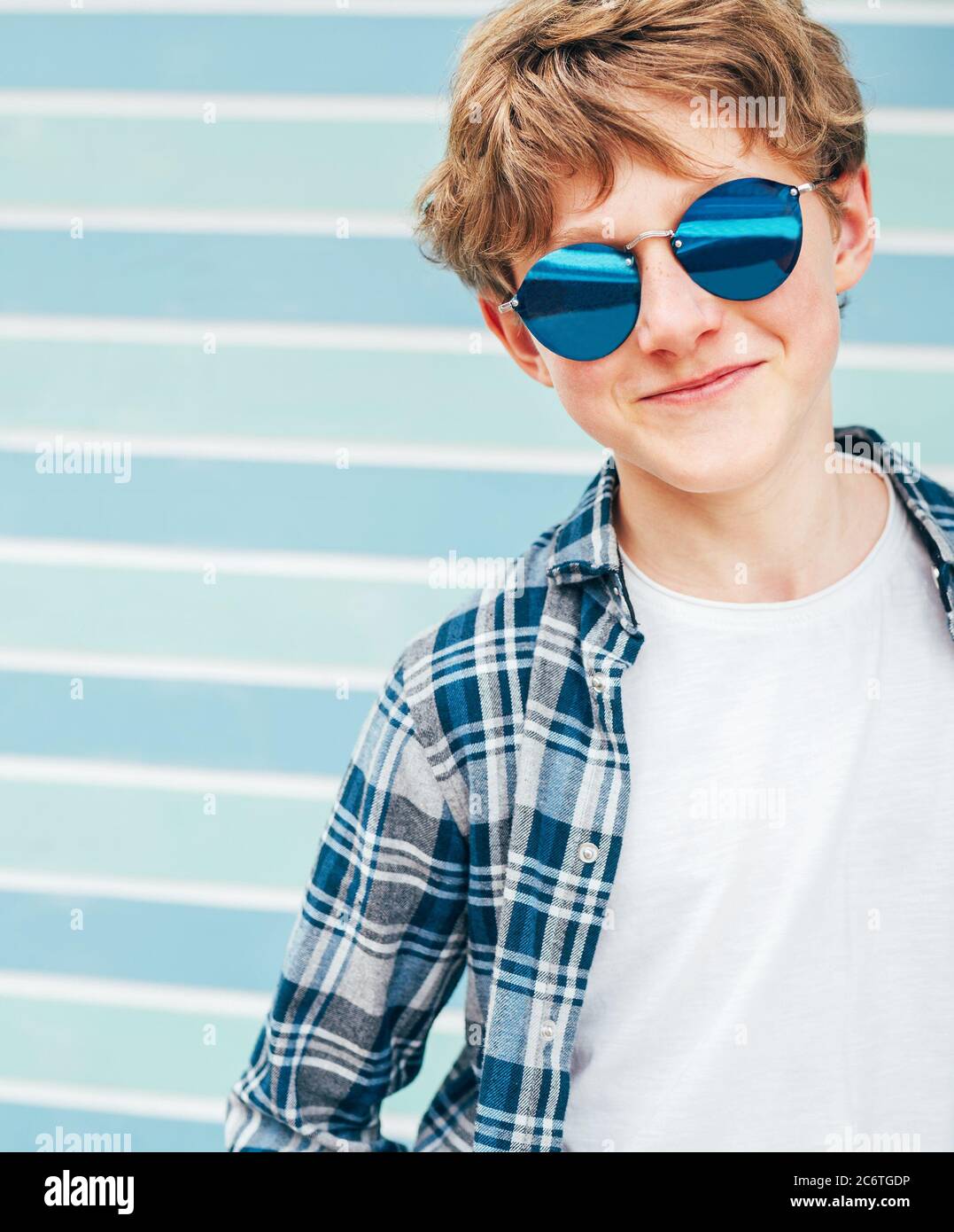 Blonde hair 12YO caucasian teenager boy Fashion portrait dressed white t-shirt with checkered shirt in blue sunglasses with turquoise blue background Stock Photo