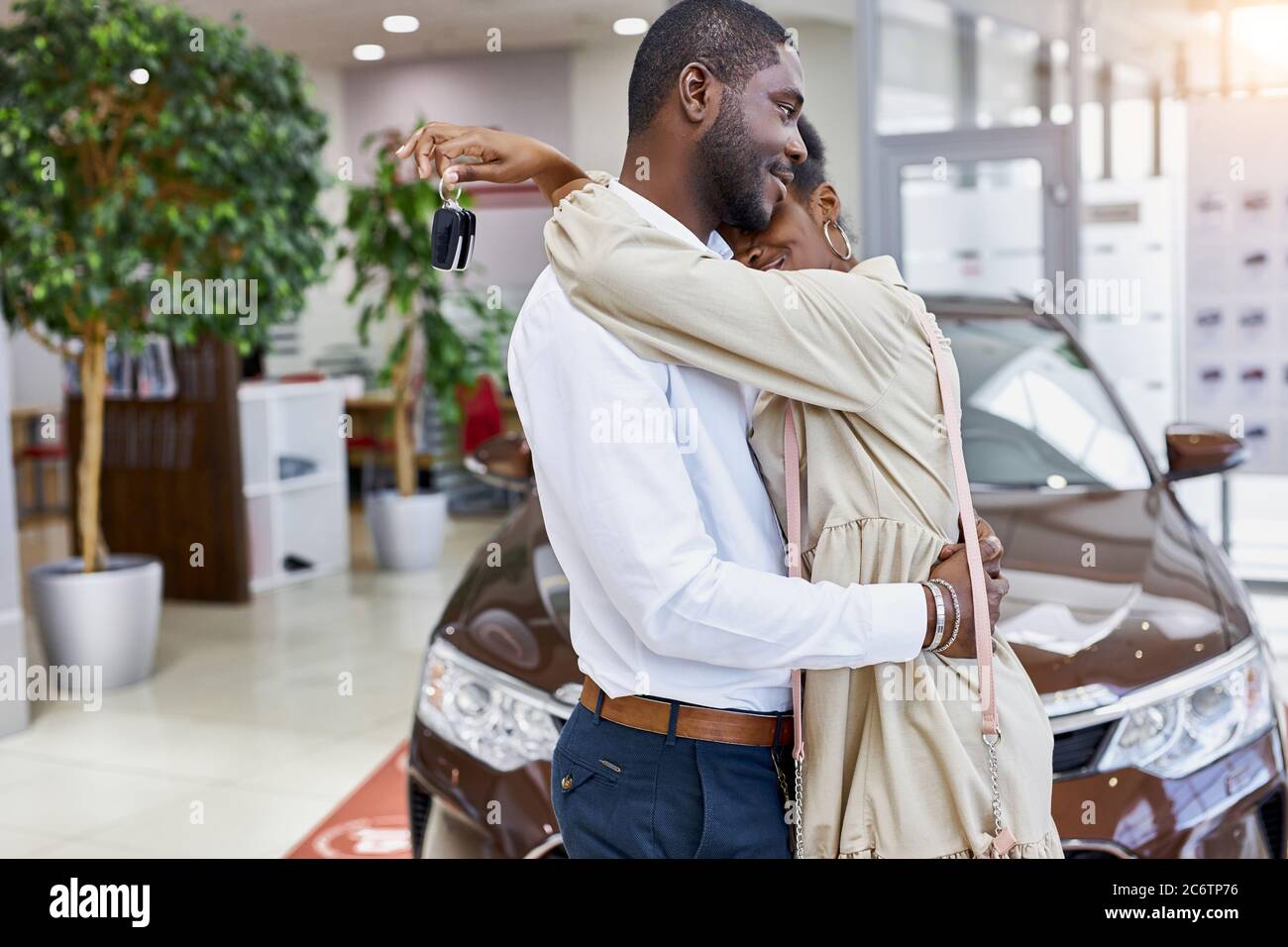 portrait of cute african married couple hugging each other in car showroom, man bought new car, woman is in love with him Stock Photo