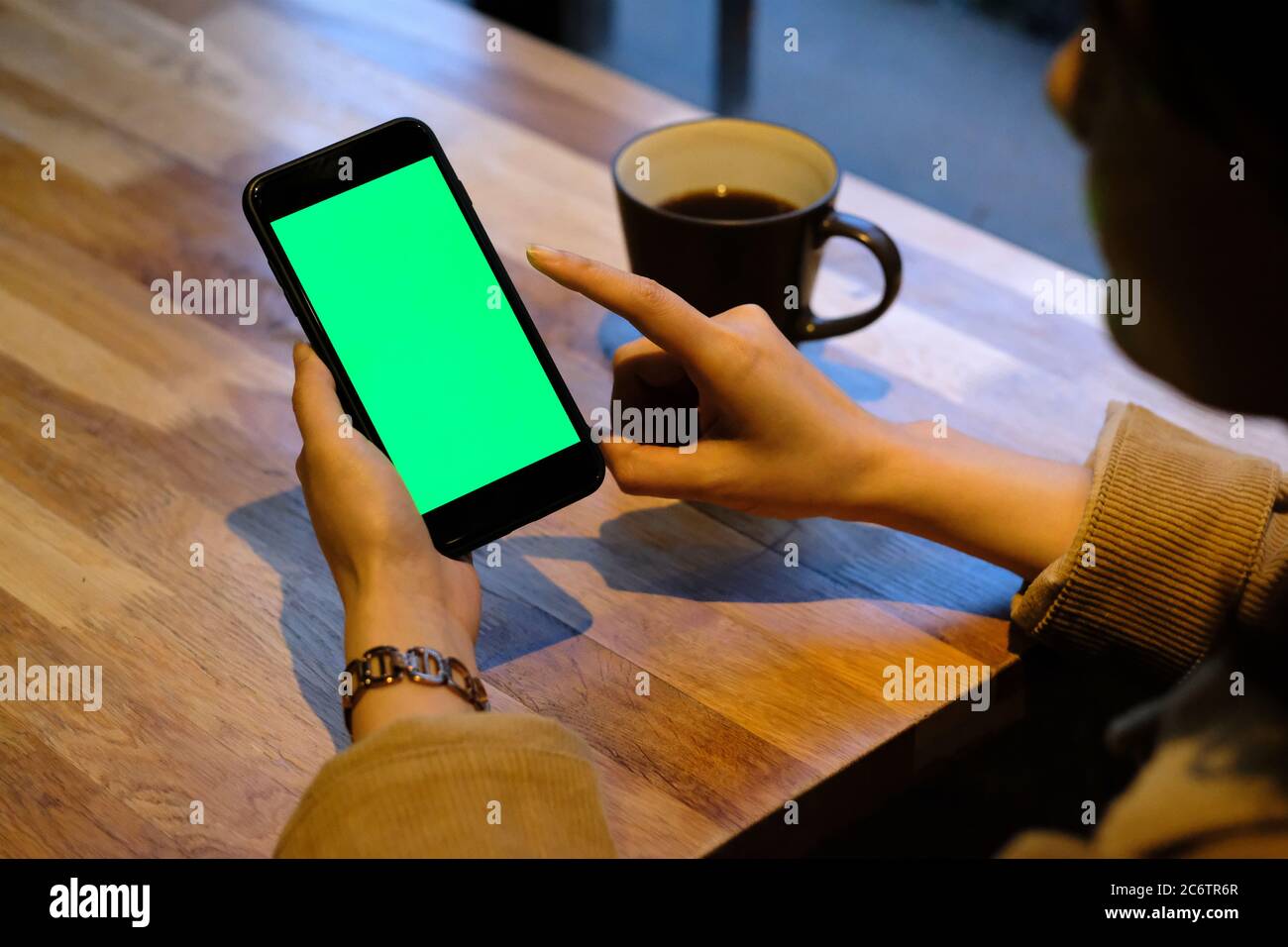 over shoulder one woman tapping green screen smartphone in cafe. A cup of coffee on wooden table. Blur background Stock Photo