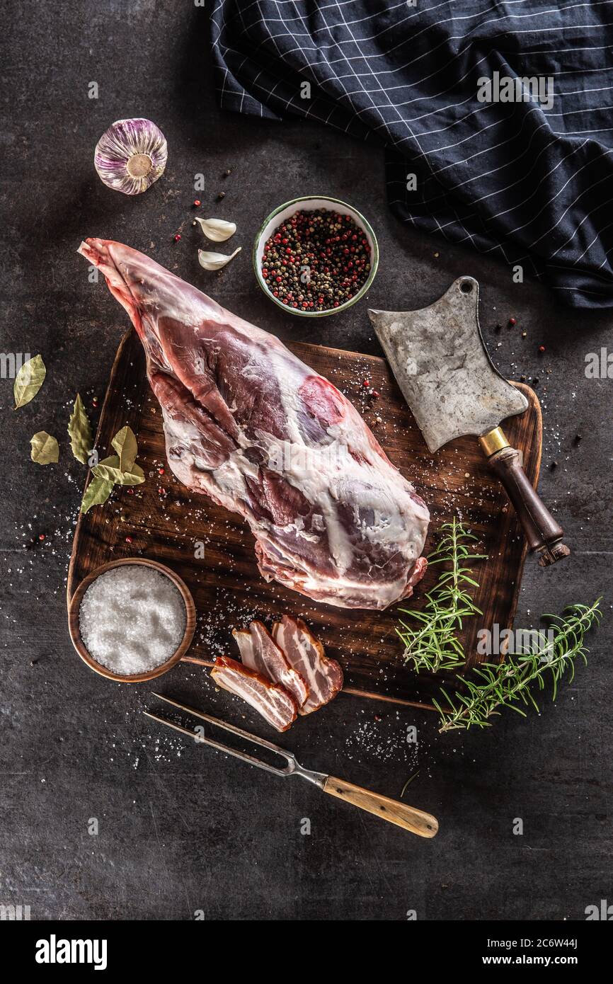 Raw lamb thigh surrounded by salt, pepper, garlic, bacon and rosemary on a vinateg surface Stock Photo