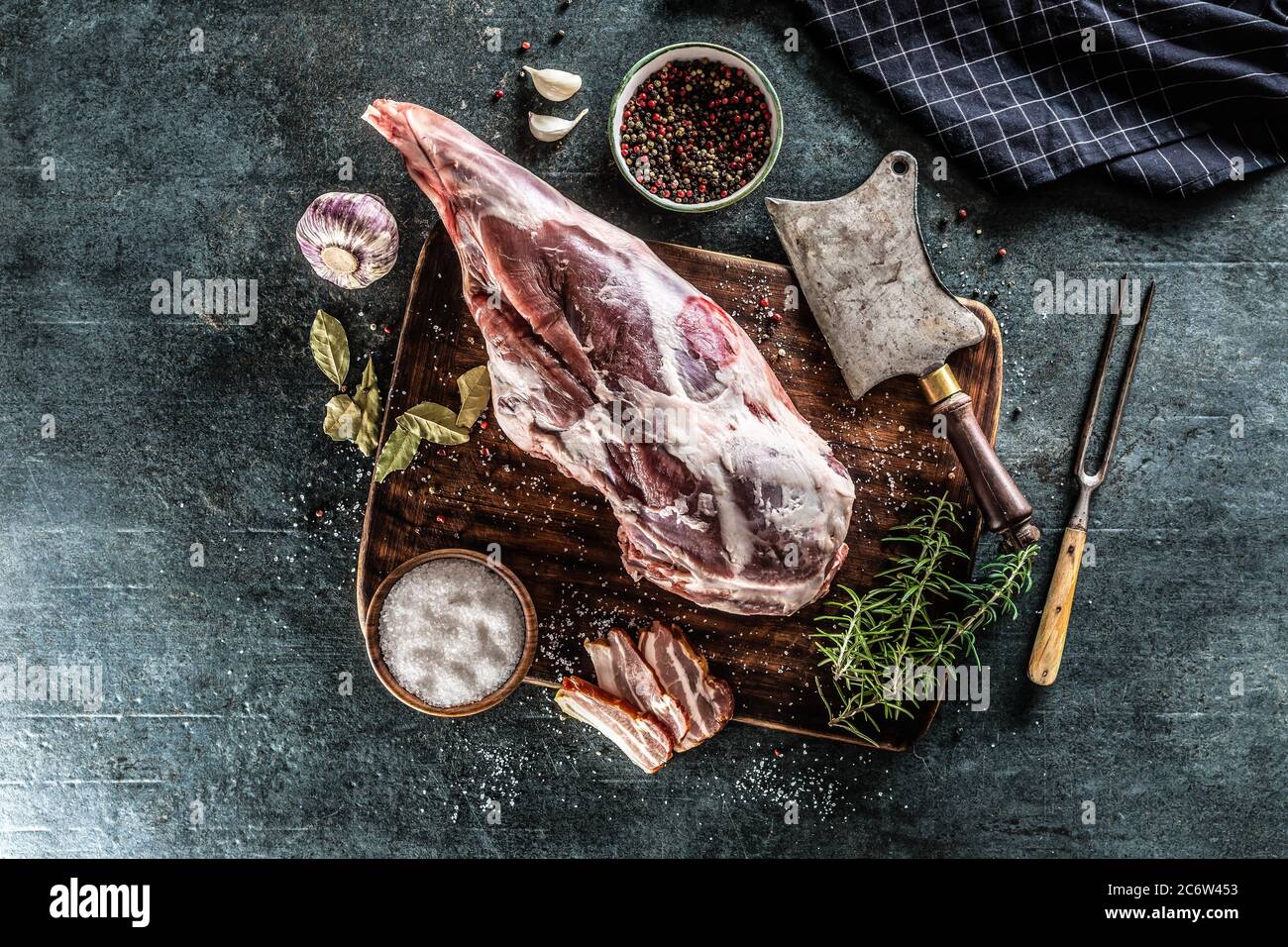 Raw lamb thigh surrounded by salt, pepper, garlic, bacon and rosemary on a vinateg surface Stock Photo