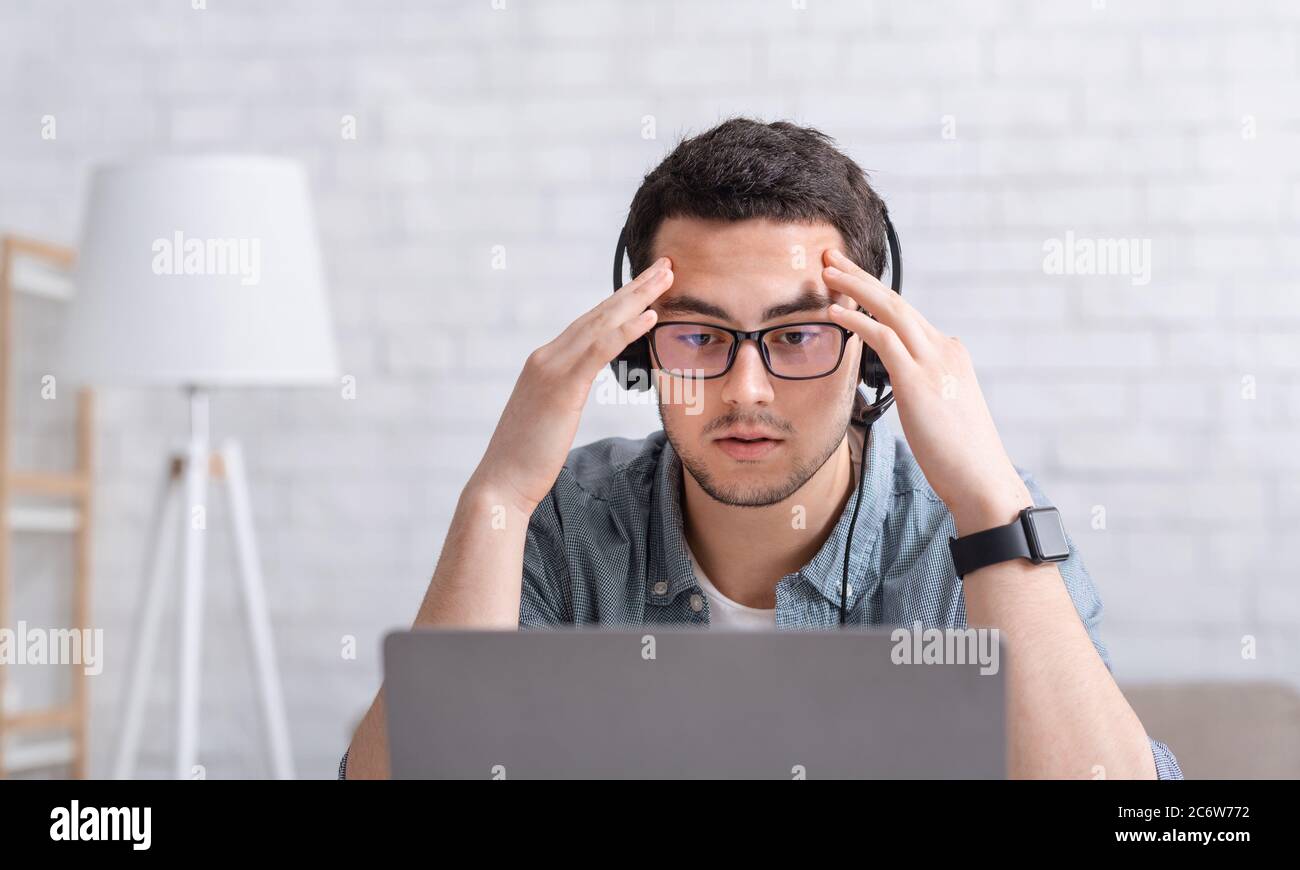 Difficult task. Concentrated guy in glasses and headphones puts hands to forehead and ponders something, looks at laptop Stock Photo