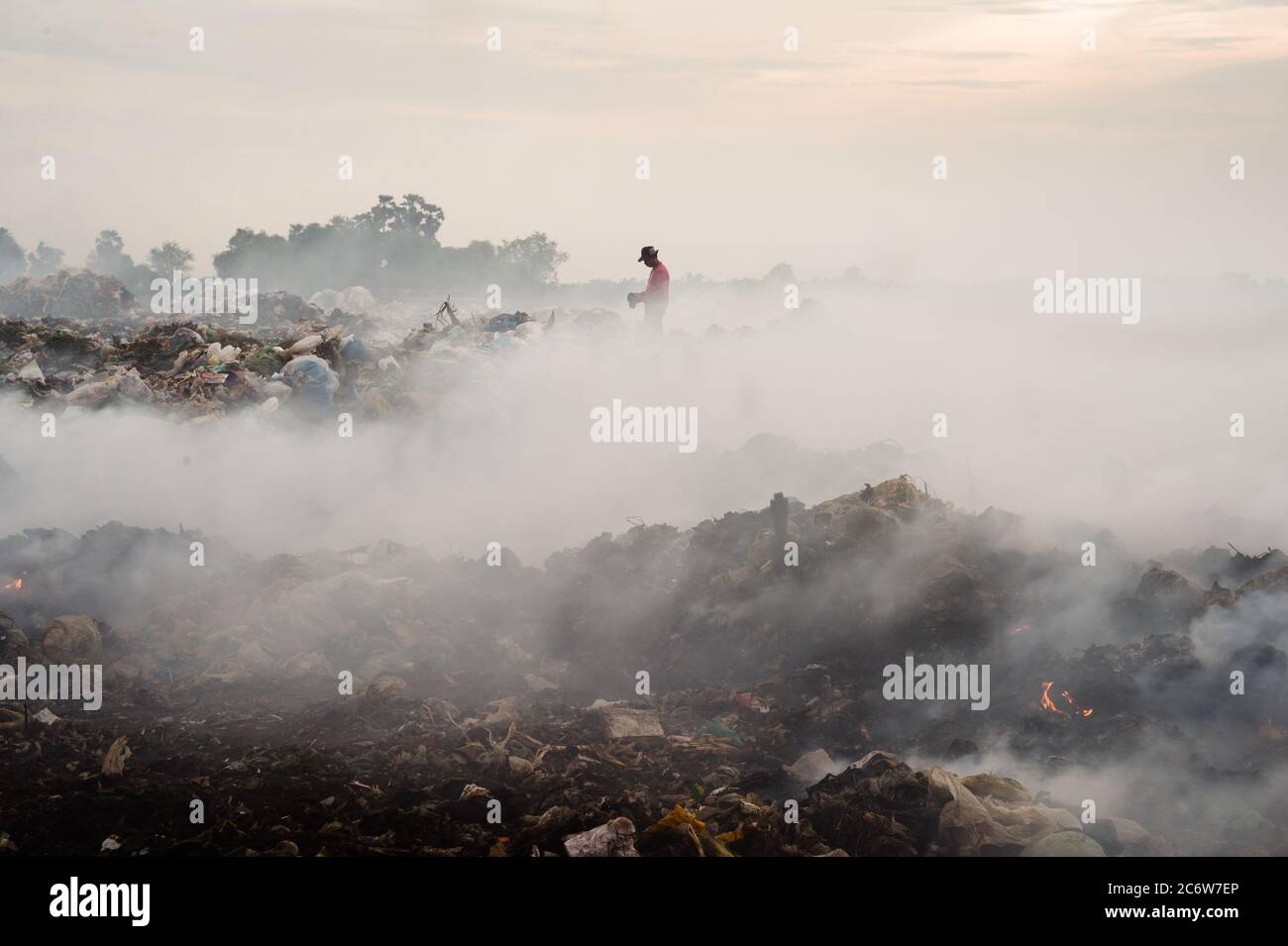 Man roams smoldering garbage dump amidst smoke Stock Photo