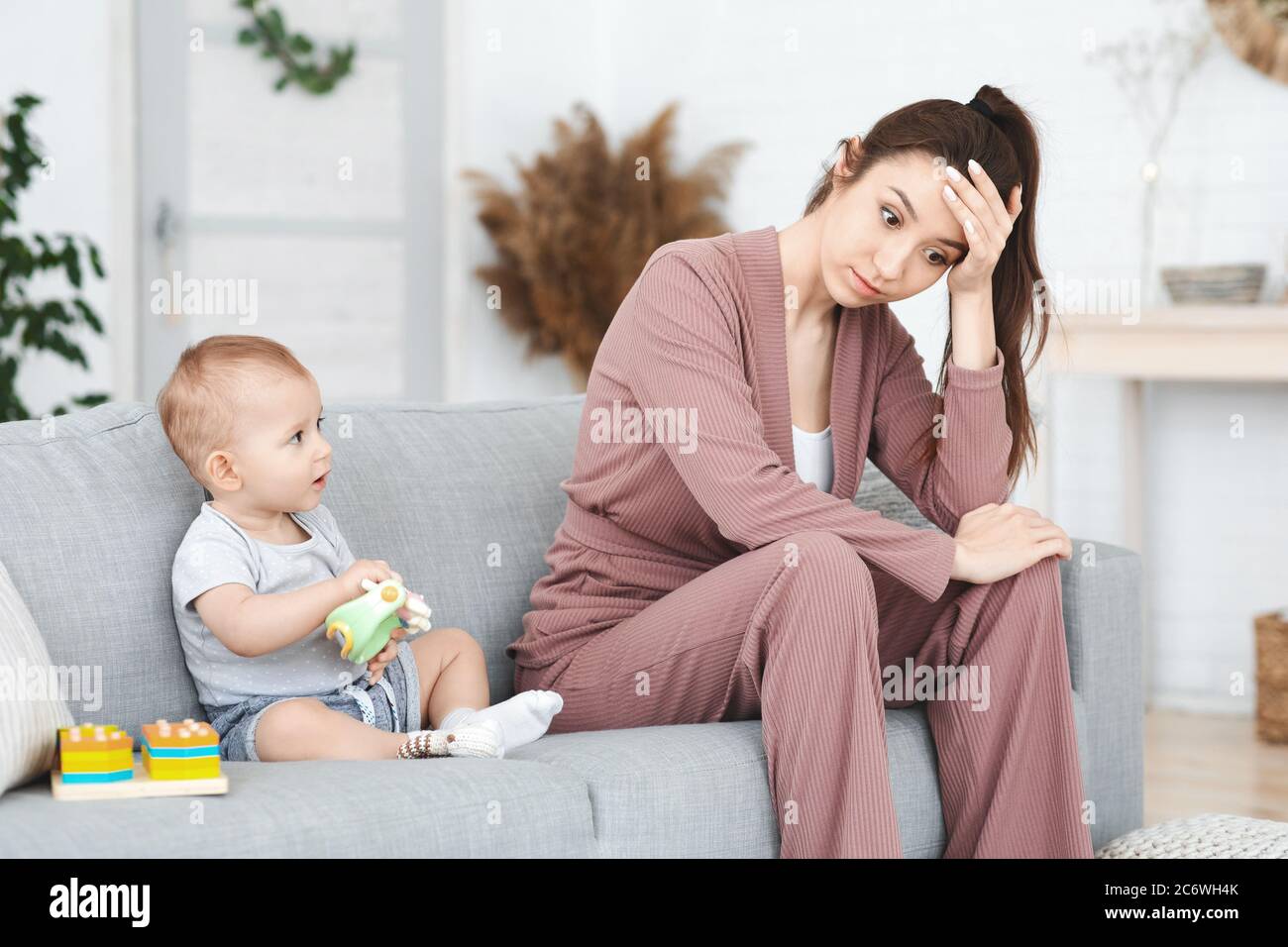 Frustration Of Motherhood. Depressed Young Woman Sitting Next To Her Toddler Baby Stock Photo