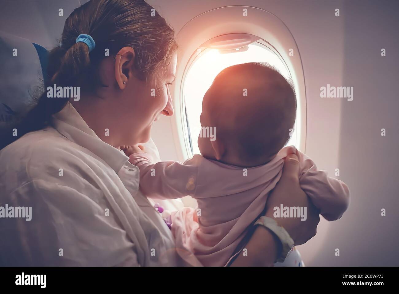 Mother with a small infant baby in her arms flying on the airplane. They look out the window with interest. Stock Photo