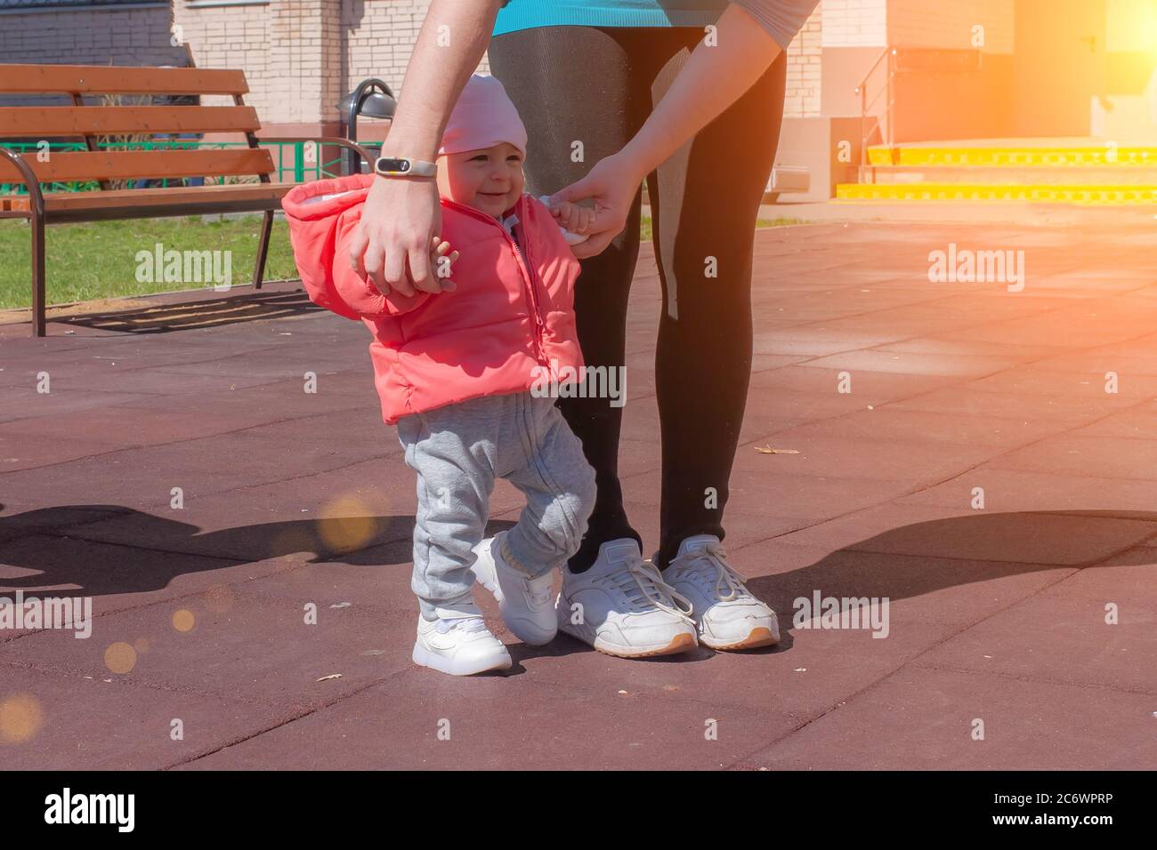 Mother holding her baby in pink by hands and teaching making first steps. Mom and infant girl walk on playground at sunny day Stock Photo