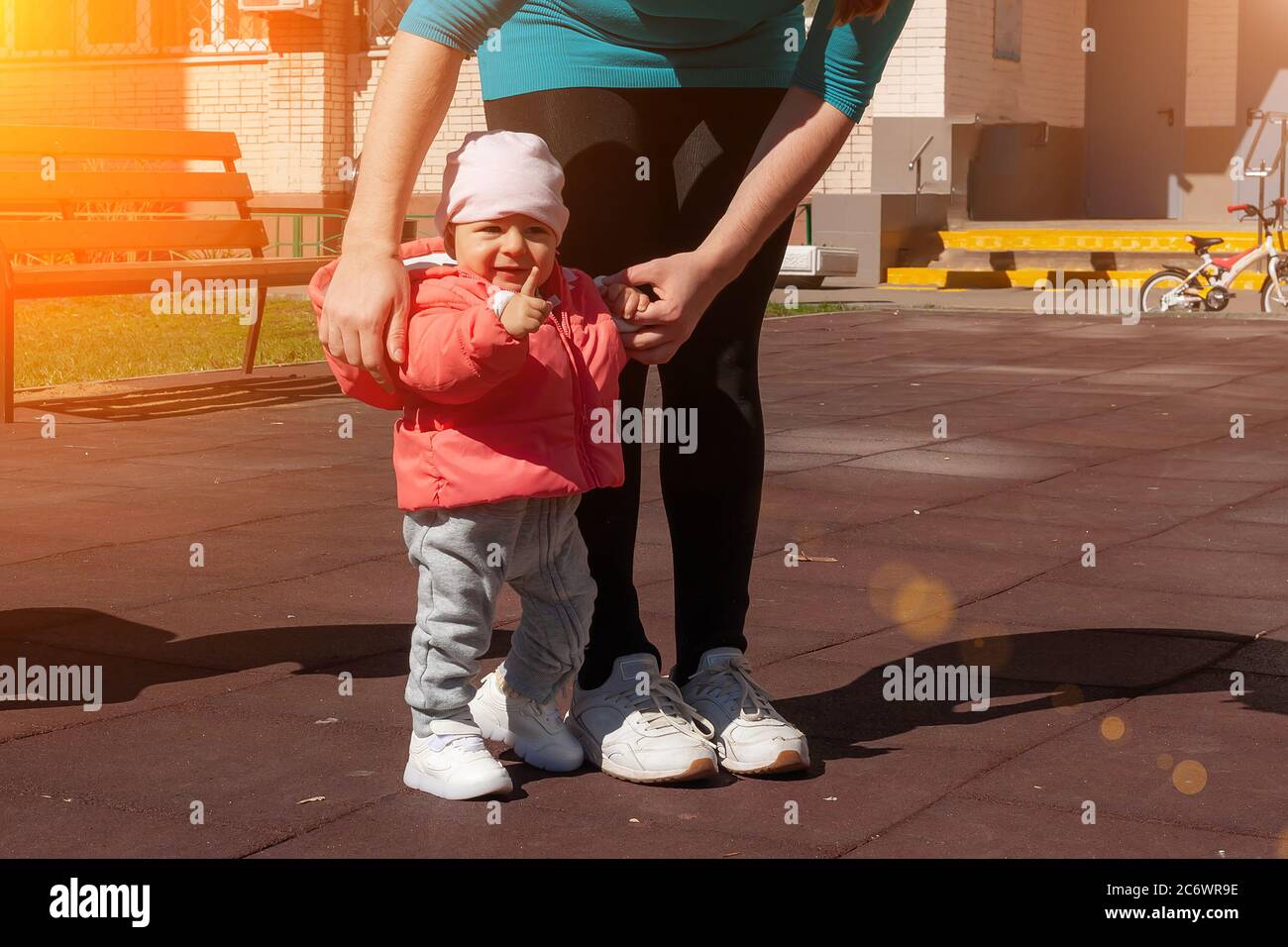 Nine month girl shows her finger. Mother holding her baby in pink by hands and teaching making first steps. Mom and infant girl walk on playground at sunny day. Stock Photo