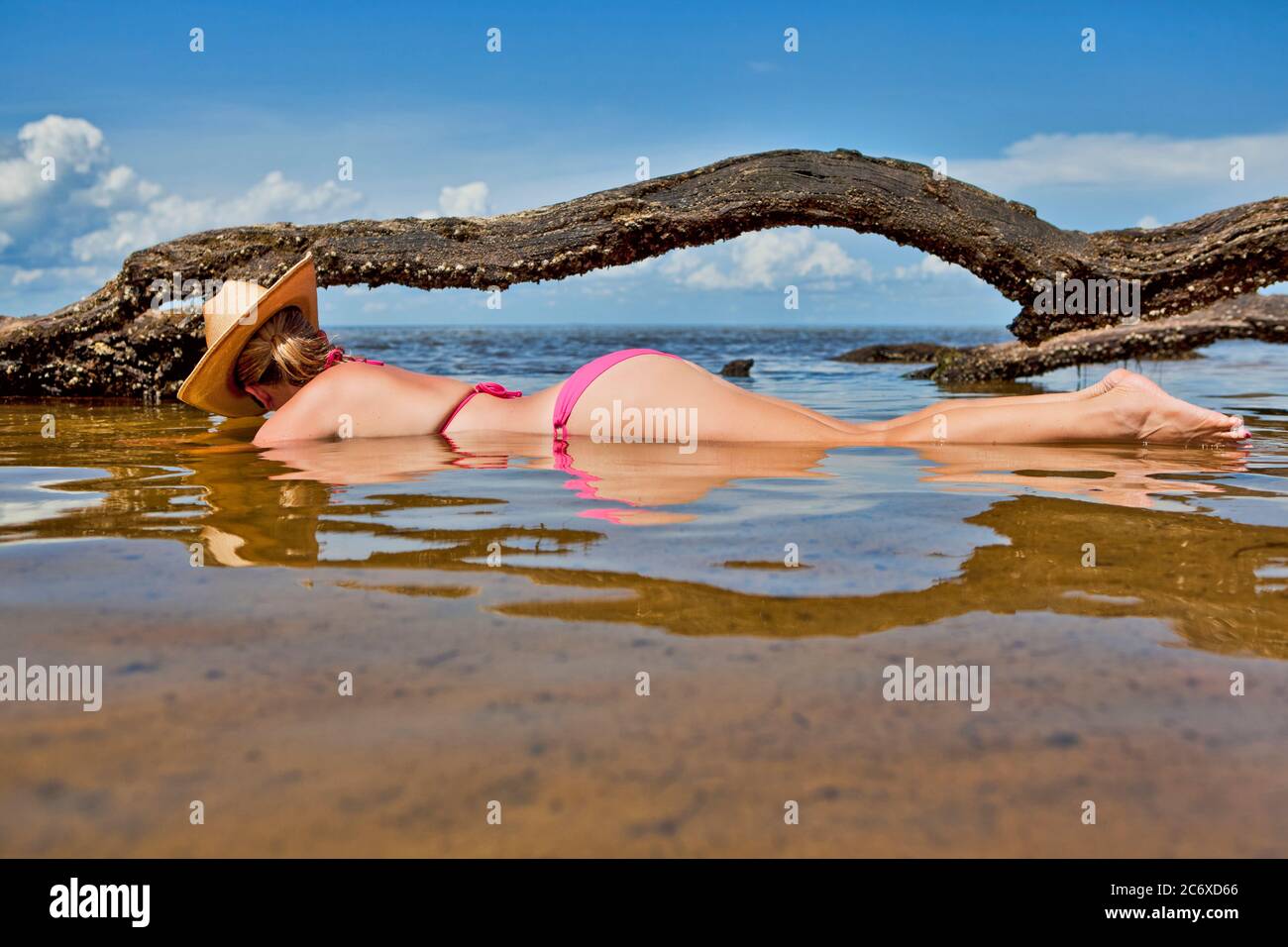 Bikini clad woman wearing a cowboy hat lying stomach down along the shoreline with driftwood behind her Stock Photo