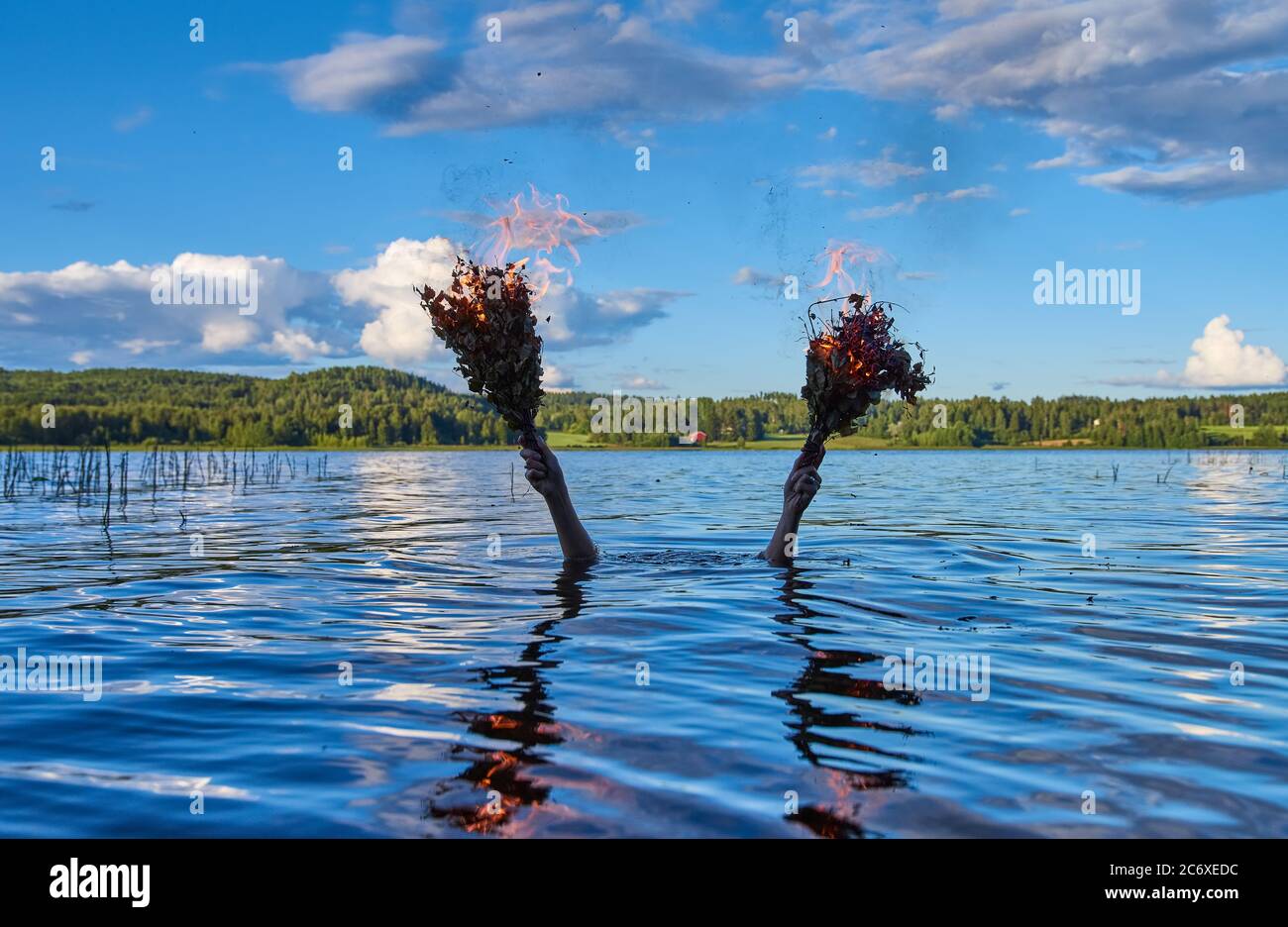 Submerged female holds two burning traditional Finnish bath whisks with her hands above water of a Finnish lake on calm summer evening. Stock Photo