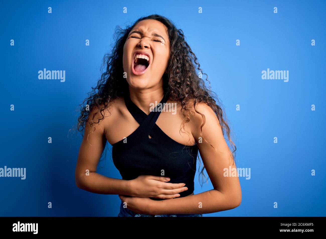 Young beautiful woman with curly hair wearing casual t-shirt standing over blue background with hand on stomach because nausea, painful disease feelin Stock Photo