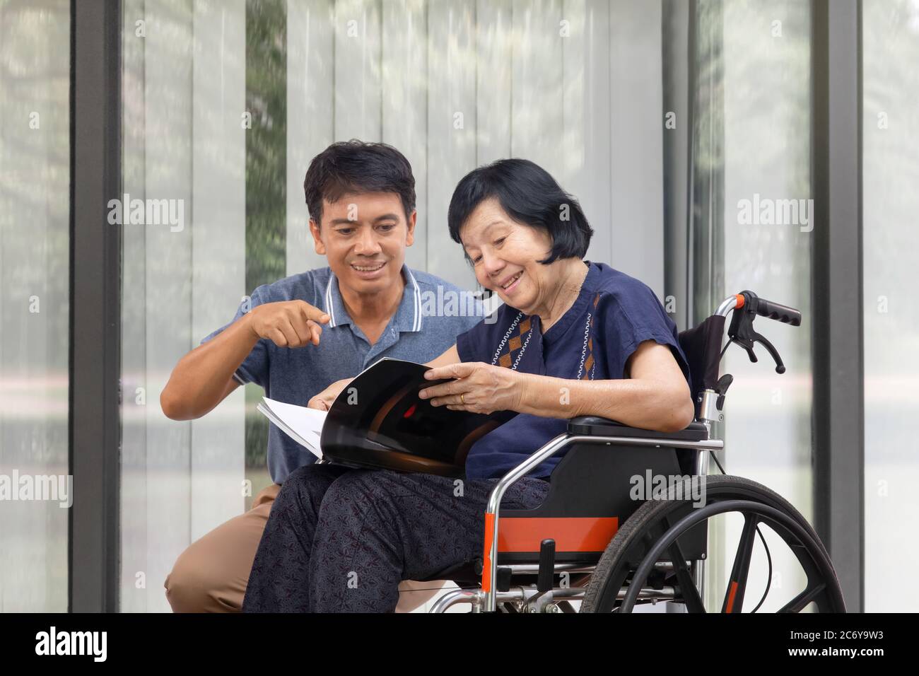 Elderly woman reading a book on wheelchair with her son take care. Stock Photo