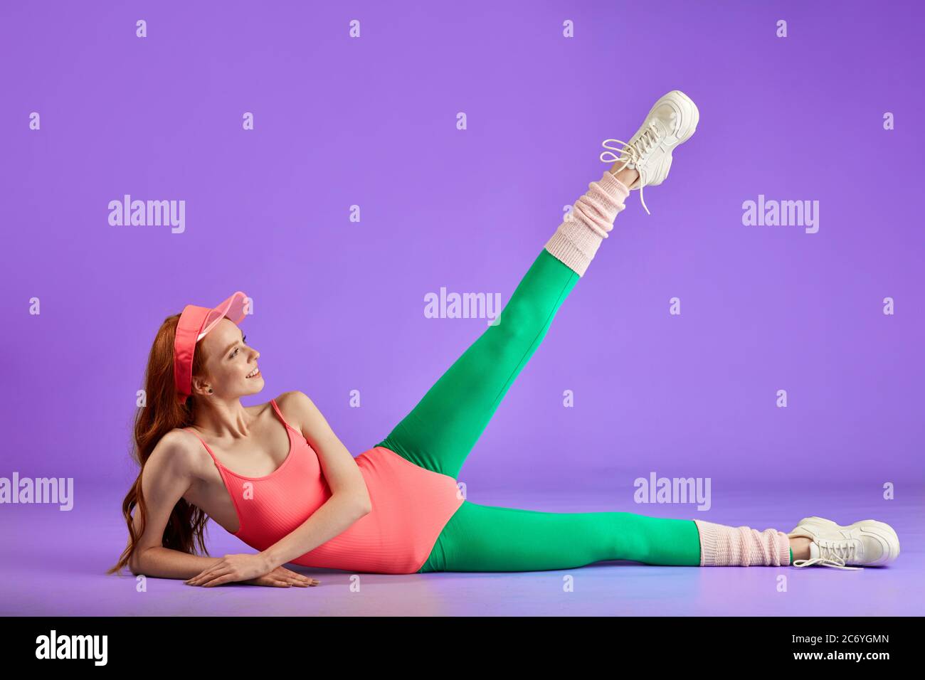aerobics female trainer shows stretching exercise, lying on the floor, lifts one leg up, wears fancy sport clothes, pink bodysuit, green leggins, sun Stock Photo