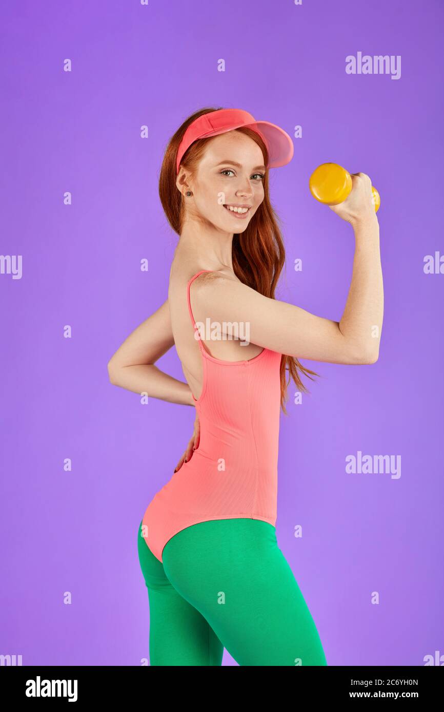 woman shoots in commercial for fitness club, works out with dumbbells, looks with pretty smile in camera, wears stylish pink bodysuite and green leggi Stock Photo