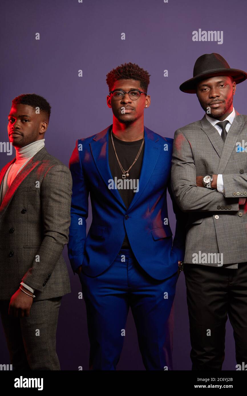 Group of three african friends dressed in stylish suits are posing isolated in studio over dark violet background. One of the guys wears hat. Classmat Stock Photo