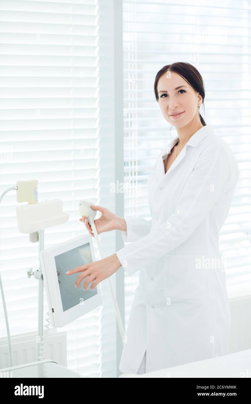 Young beautiful aestetic medicine doctor in white outfit standining near hardware cosmetology apparatus in dermatology clinic. Stock Photo