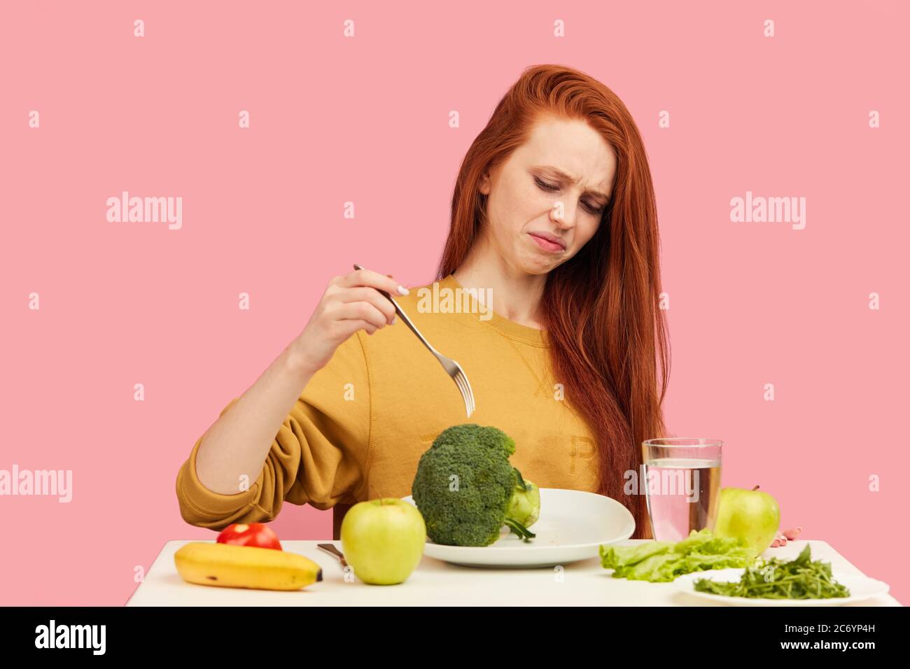 Redhead frustrated confused disgusted displeased female model makes a wry face refusing to eat broccoli and veg healthy food sitting at table over pin Stock Photo