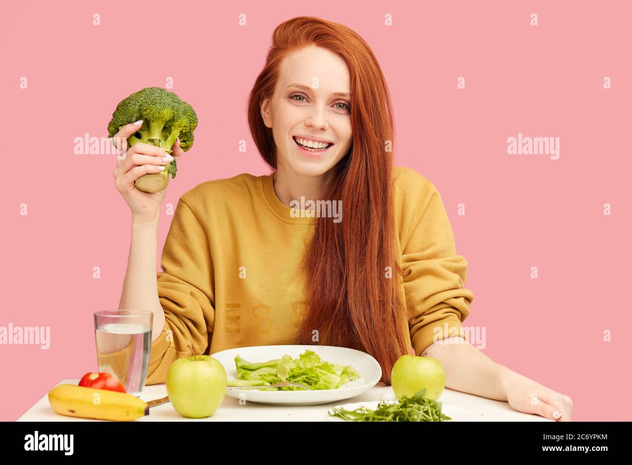 young pleasantly-looking vegg woman eating with eagerness and appetite raw green broccoli , smiling happy at camera over studio isolated pink backgrou Stock Photo