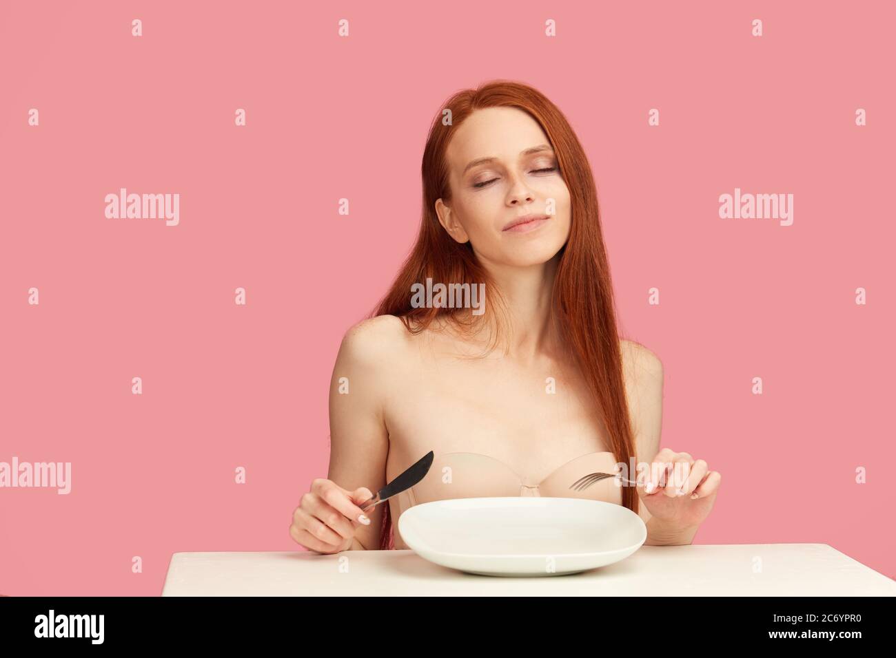 Red-haired anorexic woman dreaming about cake sitting at table with empty plate. Diet Concept. Weight Loss Problem. Starving Young Woman. Mental break Stock Photo