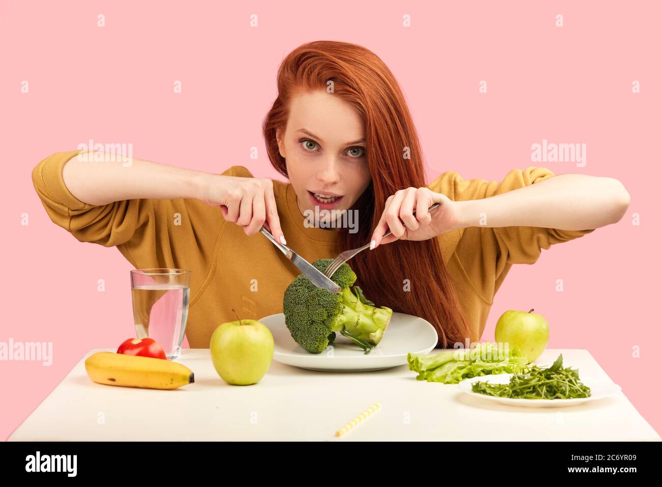 Hungry young vegg woman eating with eagerness and appetite raw green broccoli , smiling happy at camera over studio isolated pink background. Vegetabl Stock Photo