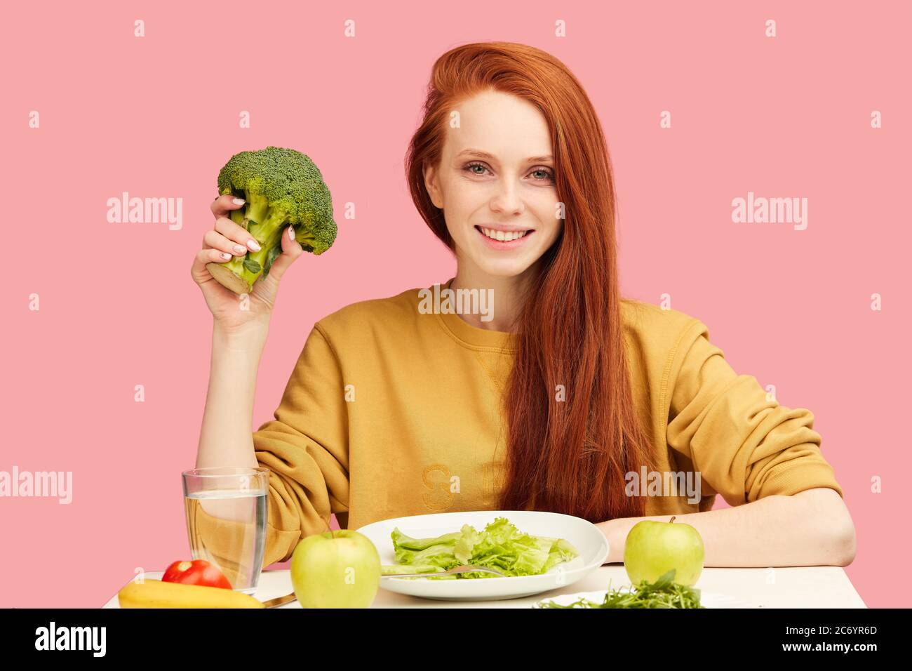 young pleasantly-looking vegg woman eating with eagerness and appetite raw green broccoli , smiling happy at camera over studio isolated pink backgrou Stock Photo