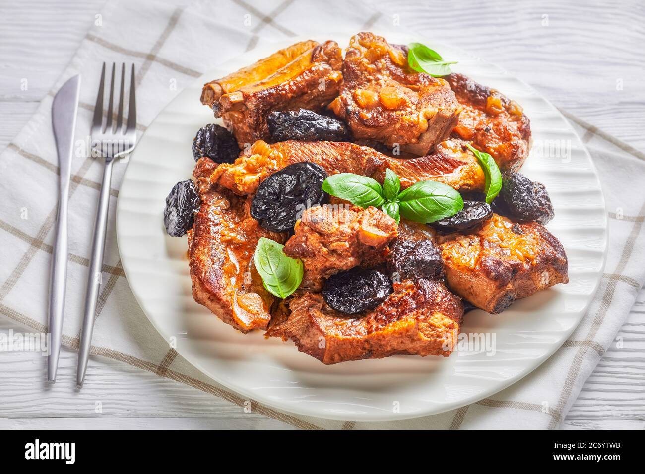 Fall recipe of short baby back bone-in pork ribs braised with prunes, broth, onion and garlic, decorated with fresh basil, served on a white plate on Stock Photo
