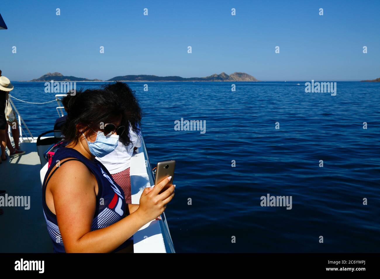 12th July 2020, Ria de Vigo, Galicia, northern Spain: A passenger wearing a face mask takes photos with her smartphone from the ferry from Vigo to the Cies Islands (in background). Spain has relaxed travel restrictions from 21st June after a strict lockdown to control the Covid 19 coronavirus and many Spaniards are returning to the beaches. The Cies Islands are a popular beach tourist destination off the coast of Galicia. Wearing face masks is still mandatory on public transport. Stock Photo