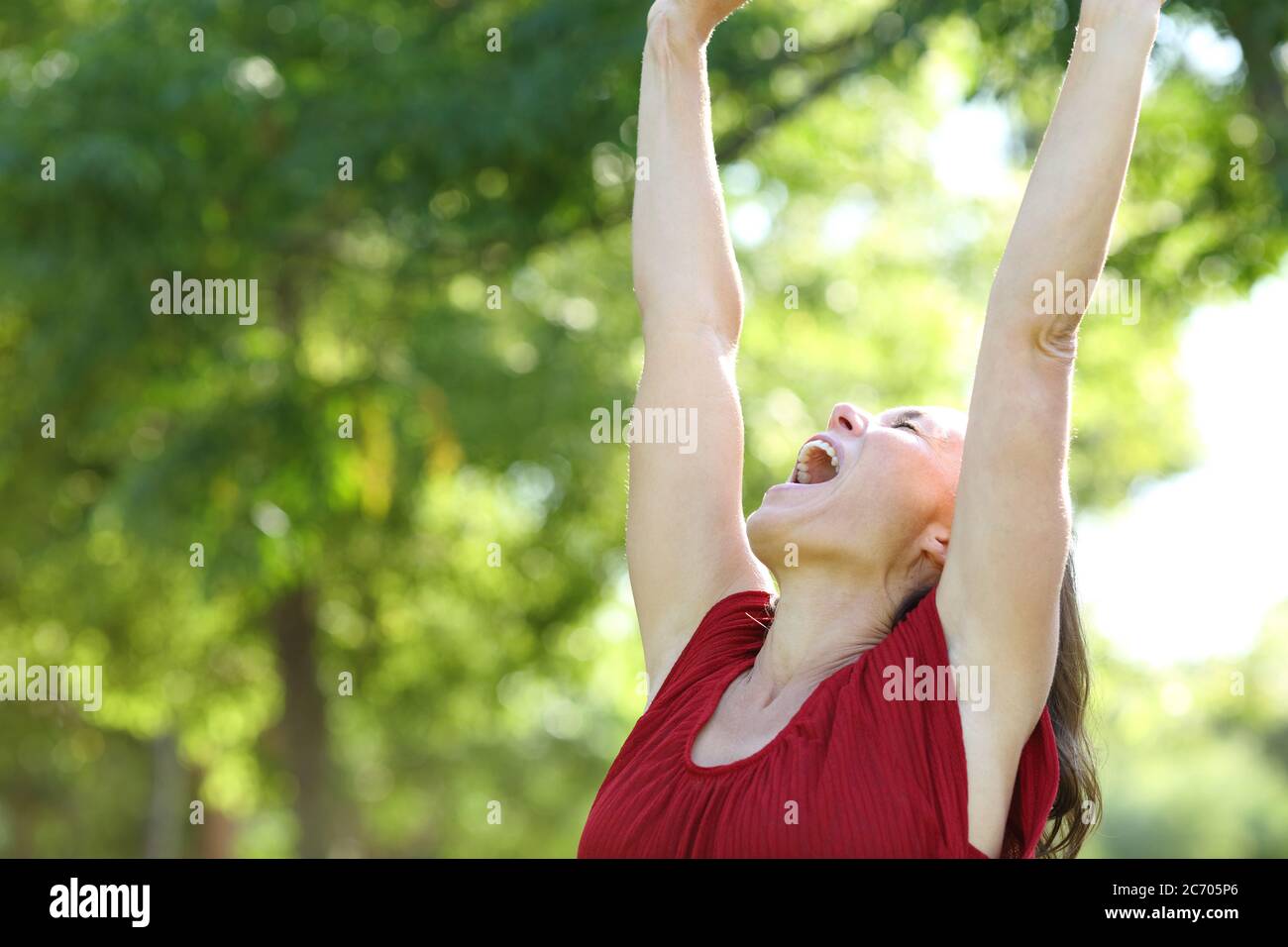 Excited adult woman celebrates raising arms standing in the park at summer Stock Photo